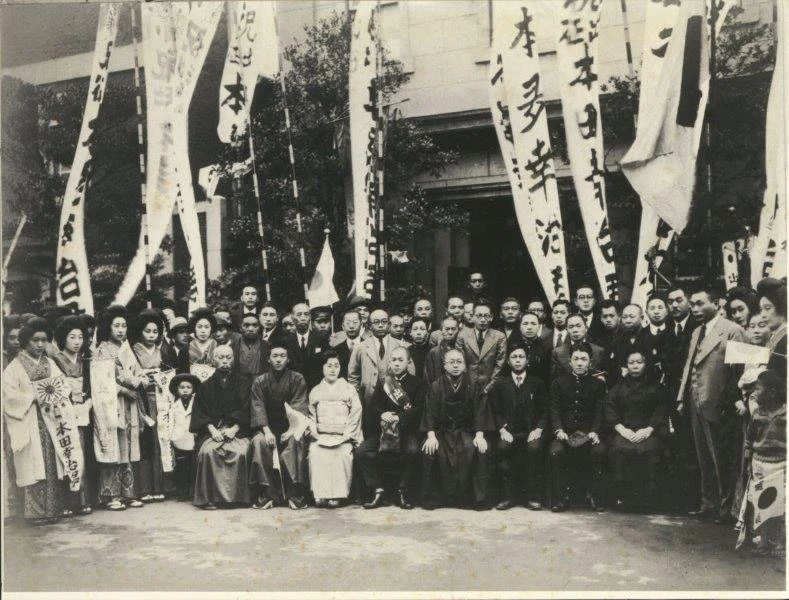 Archive photograph of a gathering outside Tsukiji Jisaku, geisha and guests assembled beneath tall congratulatory banners